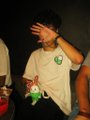 A young person wearing a white t-shirt with a logo holds their hand to their face in a dimly lit setting.