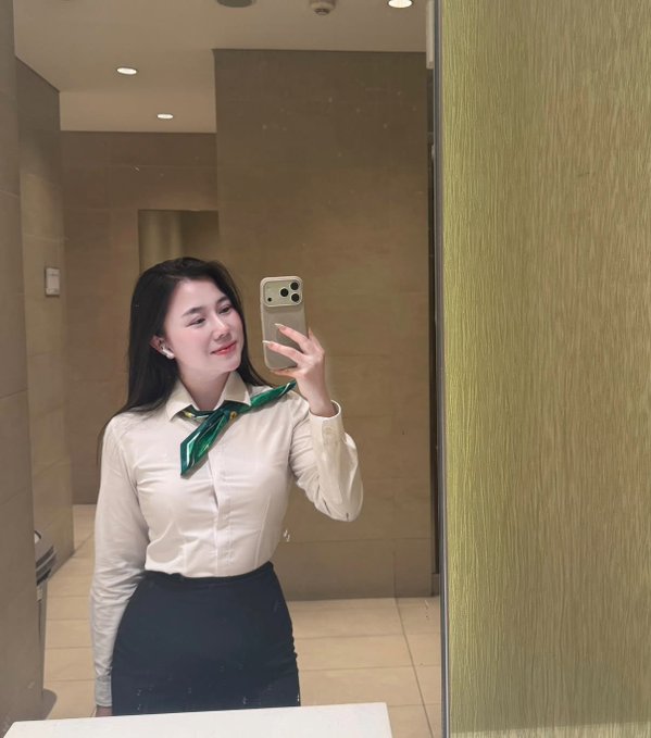 A mirror selfie of a young woman with long dark hair wearing a white button-up shirt, black skirt, and a green neck scarf, holding a smartphone in a modern bathroom.