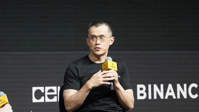 A man in a black t-shirt and glasses speaks into a yellow microphone at a panel, with a backdrop displaying the 'BINANCE' logo and partial 'cel' text.