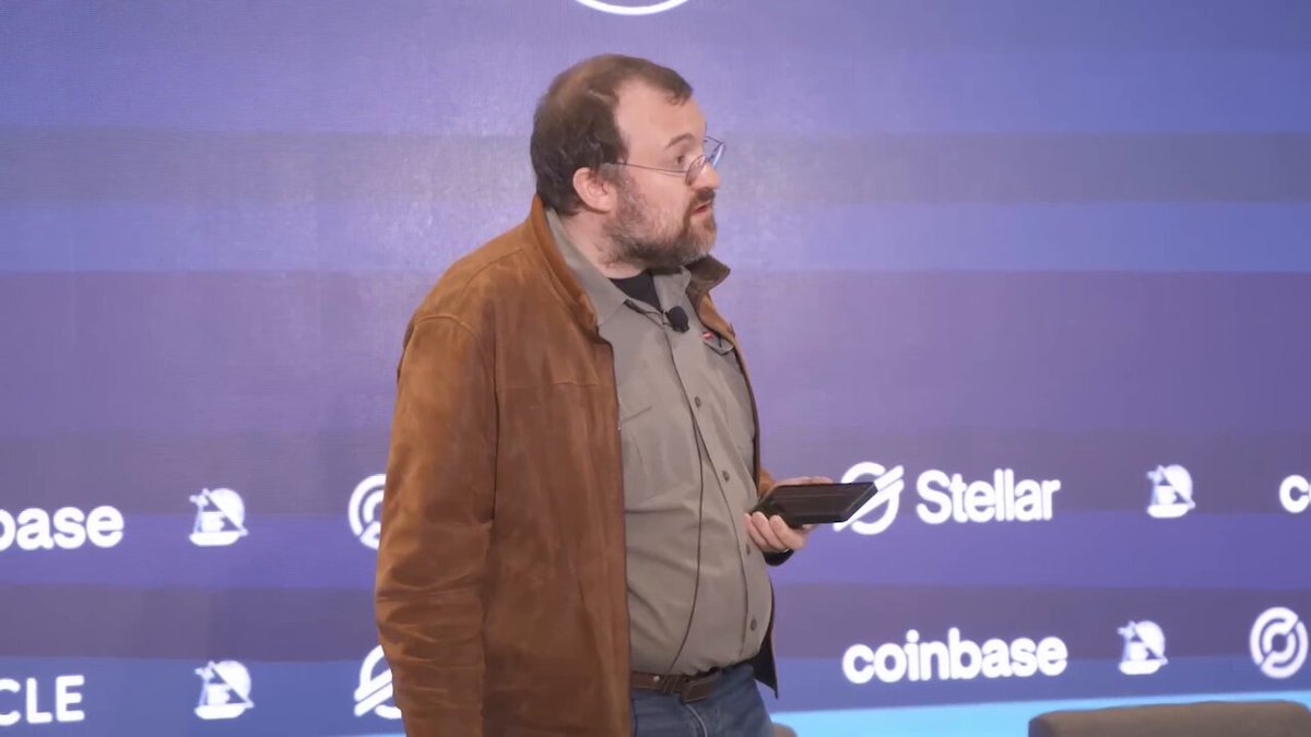 The image features a man wearing glasses standing against a blue wall. He has a brown jacket on and is holding a phone in his hand. The background includes several logos for different companies, including "Stardar", "Coinbase", and "midnight". The man appears to be engaged in a conversation or presentation related to these logos, possibly discussing the value of Bitcoin or other cryptocurrencies.