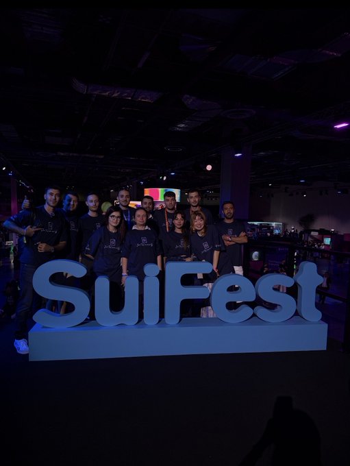 A group photo of about ten people wearing matching dark blue t-shirts posing behind a large blue 'SuiFest' sign at what appears to be a tech conference venue.