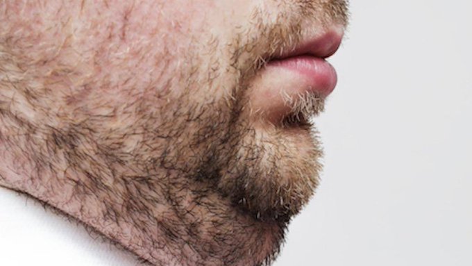 A close-up profile shot of a man's beard, mustache, and lips, showing dense facial hair against his skin.