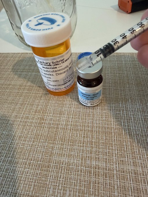 A prescription bottle of 'Zachary Schrier 100mg (2ml) Testosterone 200mg/ml Injection' next to a small vial, with a syringe drawing liquid from the vial.