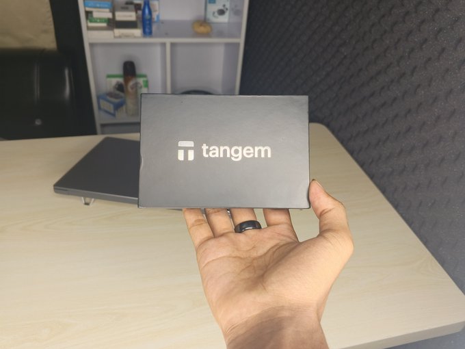 A hand holds up a gray Tangem-branded box over a wooden desk, with a ring visible on the person's finger and a blurred shelving unit in the background. The image appears to showcase a new Tangem product, likely a hardware wallet, tying in with the post teasing something new from the Tangem brand.