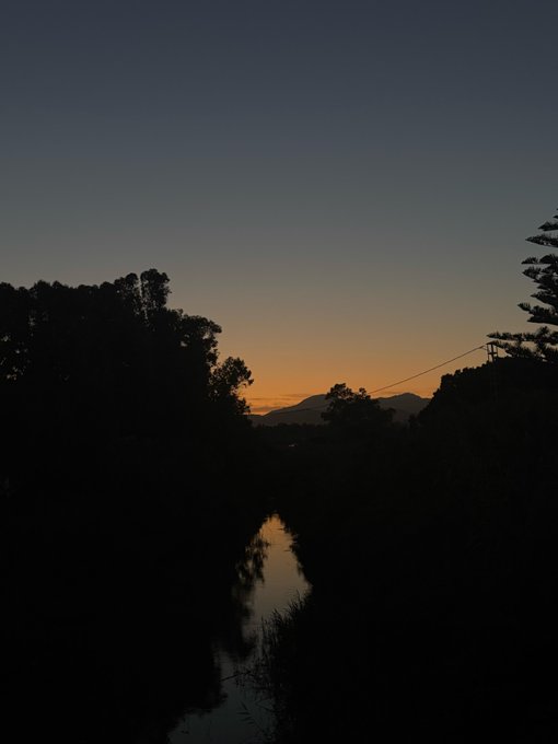 A silhouette of trees and a reflective stream against a gradient sunset sky transitioning from deep blue to warm orange near the horizon.