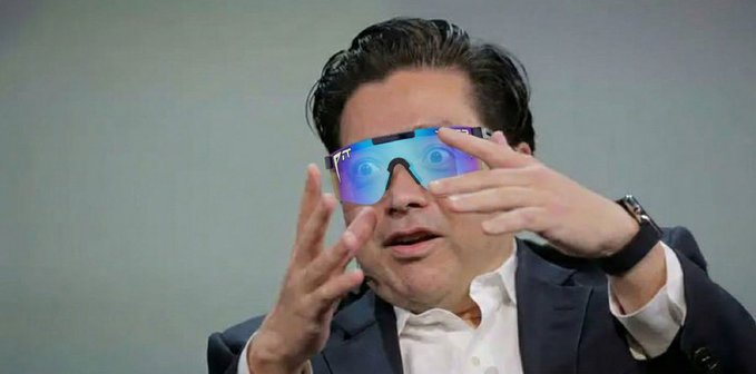 Elon Musk wears futuristic reflective blue sunglasses while adjusting them with both hands, dressed in a dark blazer and white shirt.