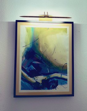 An abstract painting in blue, yellow, and white tones hangs on a wall beneath a picture light fixture.