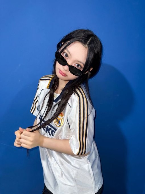 A young woman wearing dark sunglasses and a white Real Madrid soccer jersey poses against a bright blue wall.
