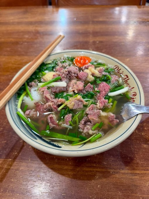 A bowl of Vietnamese pho with beef and greens, served with chopsticks and a spoon on a wooden table.