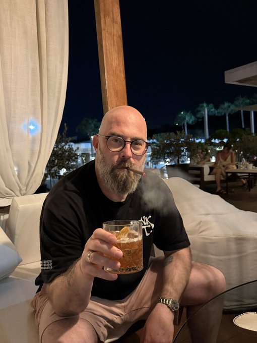 A bald, bearded man wearing glasses sitting on an outdoor lounge sofa at night, smoking a cigar and holding a glass with a drink and an orange slice.
