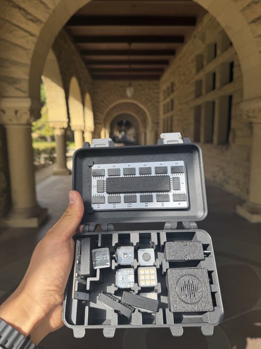A hand holds open a rugged protective case containing various small electronic devices and LED light panels, with an arched stone cloister walkway visible in the background.