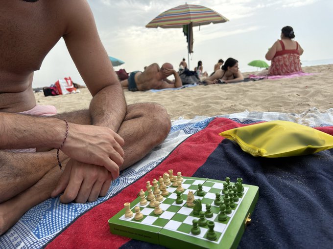 A man sits on a beach towel playing a game of chess, with other beachgoers and umbrellas visible in the background.
