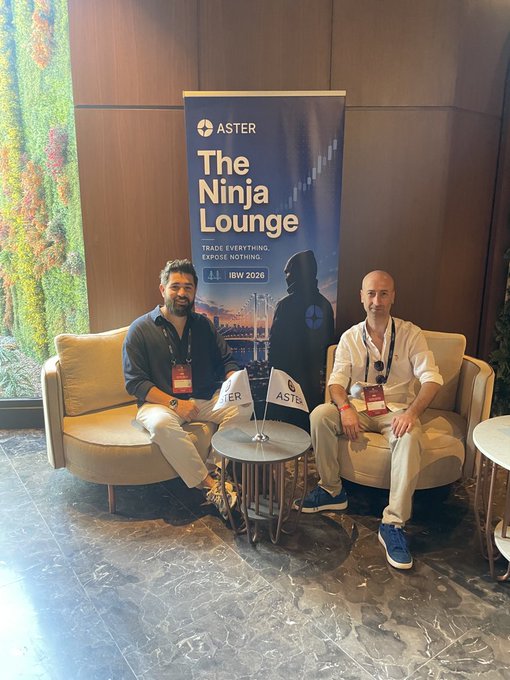 The image shows two men sitting on chairs, with a banner for "The Ninja Lounge" behind them. The banner has a blue background and white text that reads "The Ninja Lounge", indicating an event or gathering related to the ninja theme. The men are seated in front of a table, which is placed against a wall adorned with a painting.