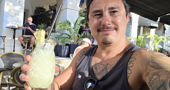 A tattooed man in a black tank top smiles while holding a pineapple-shaped cocktail glass at an outdoor cafe.