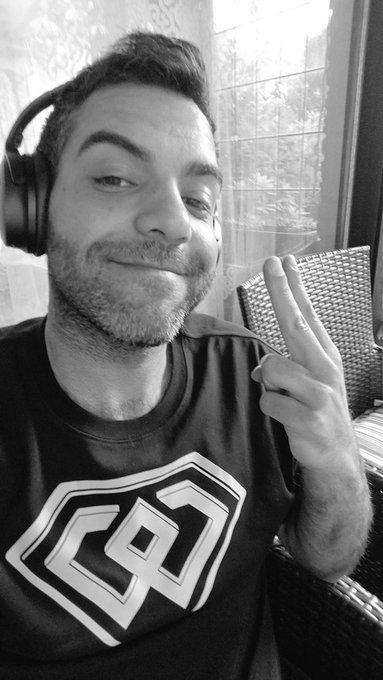 A black and white selfie shows a man with headphones smiling, making a peace sign while holding a leaf, wearing a black t-shirt with a white logo.