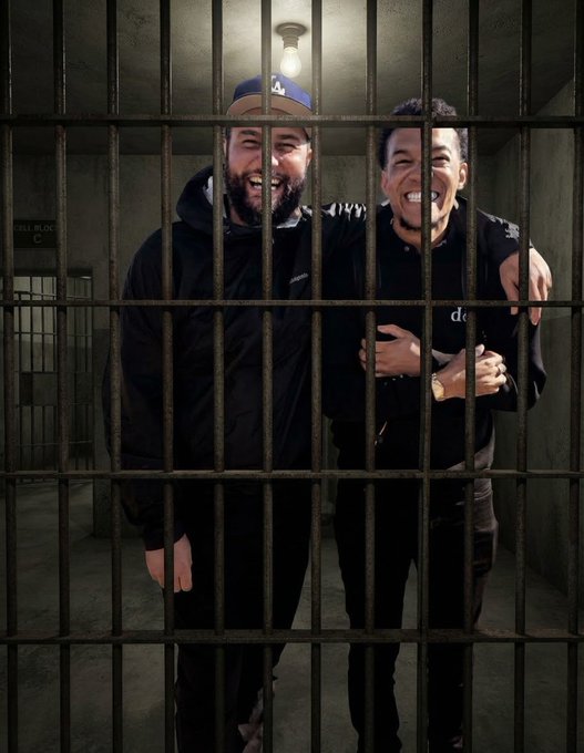 Two men standing behind prison bars in a dimly lit cell, laughing together with their arms around each other.