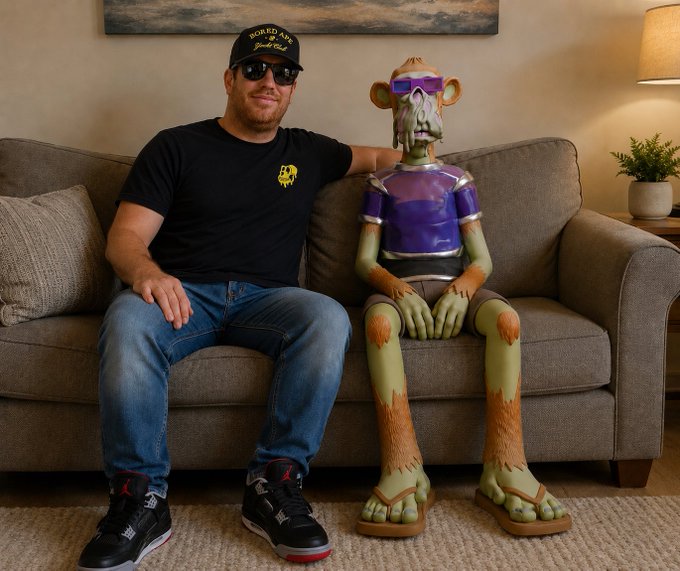 The image features a man sitting on a couch with a large, green, and purple statue of a monkey or gorilla behind him. The statue appears to be made out of foam or plastic and has a pair of sunglasses on its face. The caption "GM You are never alone when you have an ape" is written above the statue, which seems to be related to the man's interaction with the large animal figure.

The man in the image is wearing a black t-shirt and blue jeans while sitting on the couch. There is also a potted plant visible near the couch, adding some greenery to the scene.