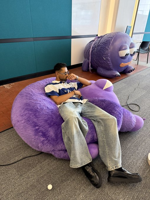 A man lounges on a giant purple Yeti mascot plush, with another purple creature sculpture in the background, fitting the playful "infiltrated the Monad office" vibe of the post.