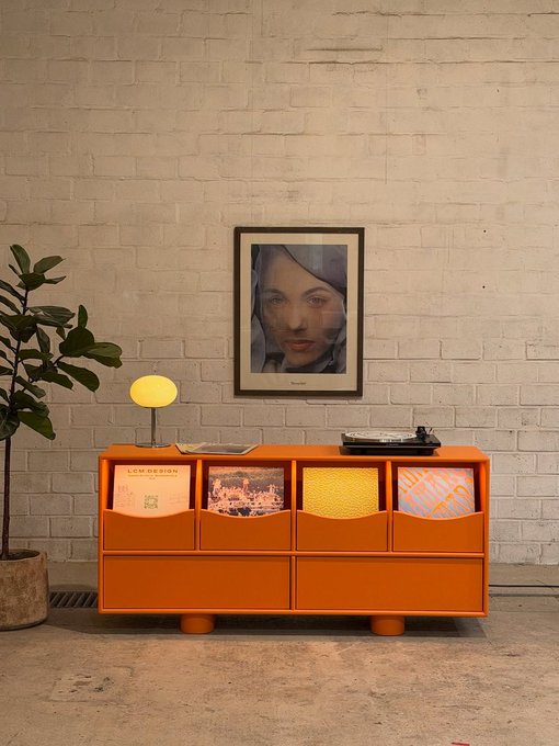 An orange record storage cabinet labeled 'LOW DESIGN' holding various vinyl records, with a framed portrait of a woman on the white brick wall above and a turntable on top.