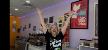 A person celebrating with arms raised inside what appears to be a smoothie shop, with a sign reading '3 DAYS TO PROMO CHAOS' visible in the background.