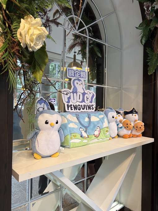A decorative display featuring plush penguin toys and a tote bag arranged on a white shelf, with a neon sign reading 'PUPU PENGUINS' and Chinese characters '胖企鵝' in the background.