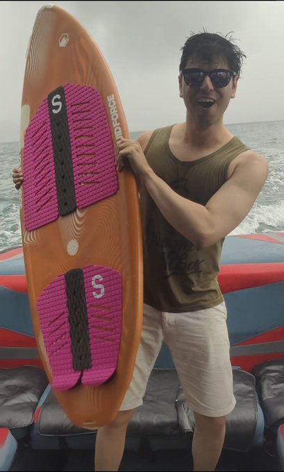 A man in sunglasses and a green tank top smiles on a boat while holding an orange surfboard labeled "SURF FORGE" with a pink "S" footpad, fitting the post's celebratory caption about turning a year older and enjoying a memorable week in Hong Kong that included wakesurfing.