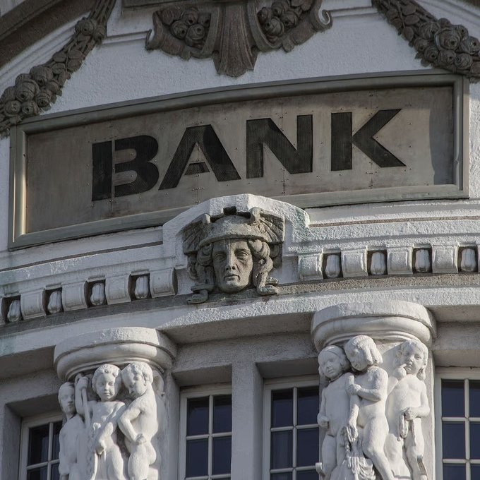 The ornate stone facade of an old building featuring the word 'BANK' carved above decorative carved figures and a sculpted face.
