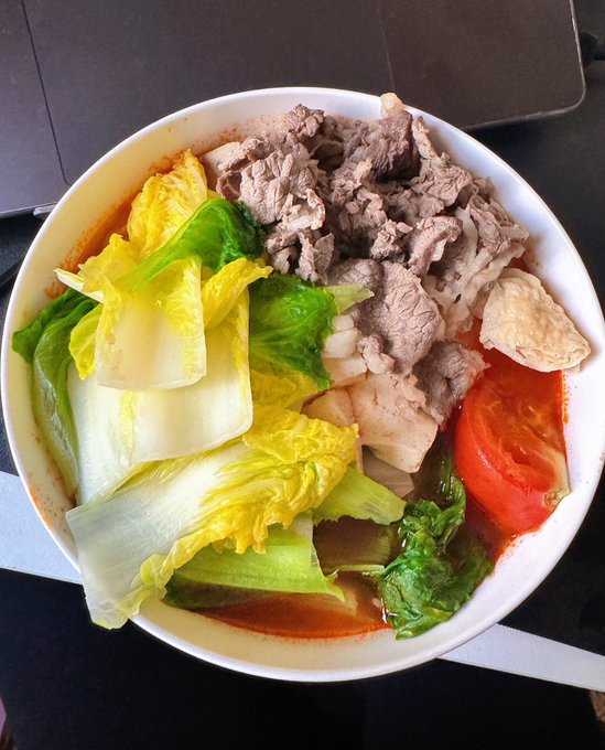 Overhead view of a white bowl filled with hot pot containing sliced beef, napa cabbage, bok choy, tomato, and other vegetables in a reddish broth.