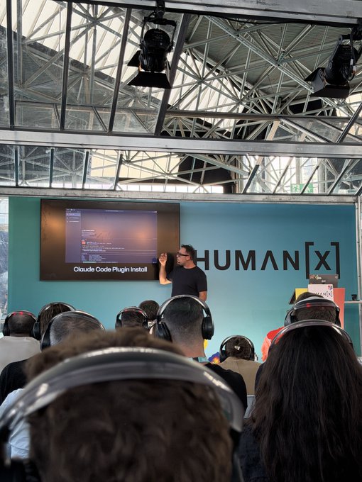 A presenter at Human[X] Amsterdam demonstrates installing the Claude Code Plugin on stage, with a slide reading "Claude Code Plugin Install" visible on the screen behind him as the audience—many wearing translation headsets—watches attentively. The talk illustrates Walrus Memory's solution to switching LLMs without losing shared history and context.