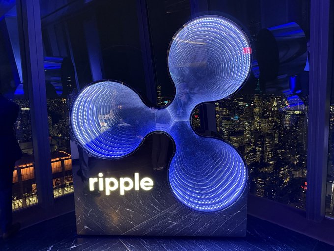 An illuminated Ripple logo display featuring the iconic three-circle cryptocurrency symbol in blue LED lights, set against a nighttime city skyline backdrop.