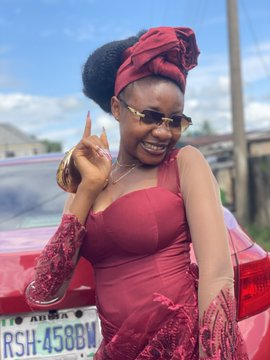 A smiling woman wearing a maroon dress, sunglasses, and a matching headwrap makes a peace sign in front of a car with a license plate reading 'RSH-458BW'.