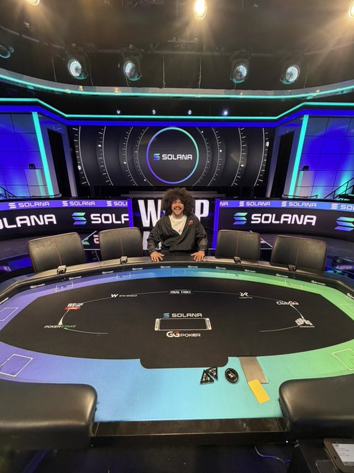A person with curly hair posing playfully behind a poker table on a brightly lit stage, with prominent 'SOLANA' branding displayed on screens and banners in the background.