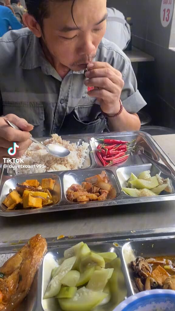The image shows a man sitting down with a tray of food in front of him. The tray contains various dishes such as rice, vegetables, and meat. The man appears to be eating from one of these dishes while looking at his phone. There is also a caption below the photo that reads "Cool weekend afternoon, my loves, whichever one you pick Time to keep grinding @wallchain !" It seems like there might have been an interaction or conversation between the man and someone else through the post.