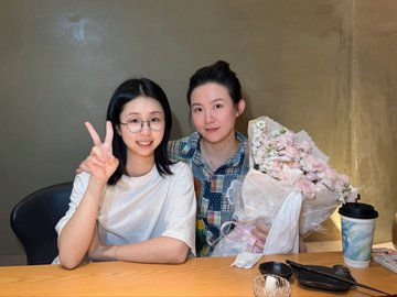 Two women pose closely together at a wooden table, with the woman on the left flashing a peace sign and wearing glasses, while the other holds a bouquet of cotton candy-colored flowers. The image accompanies a heartfelt post about a cherished friend tagged @kajiweb3, commemorating their first real photo together and the meaningful gifts—like a backpack set—she's given.