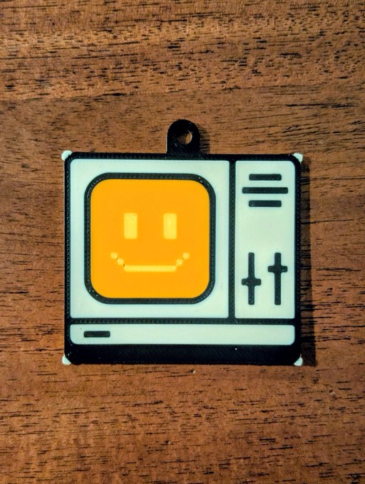 A small rubber charm or keychain depicting a retro-style television with a smiling face on its orange screen, resting on a wooden surface.