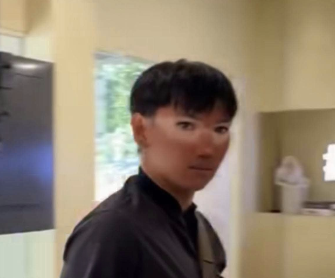 A man with short black hair wearing a dark jacket looks directly at the camera with a serious, slightly suspicious expression as he glances back over his shoulder while entering a room, perfectly capturing the awkward anticipation of returning to work after an extended absence.
