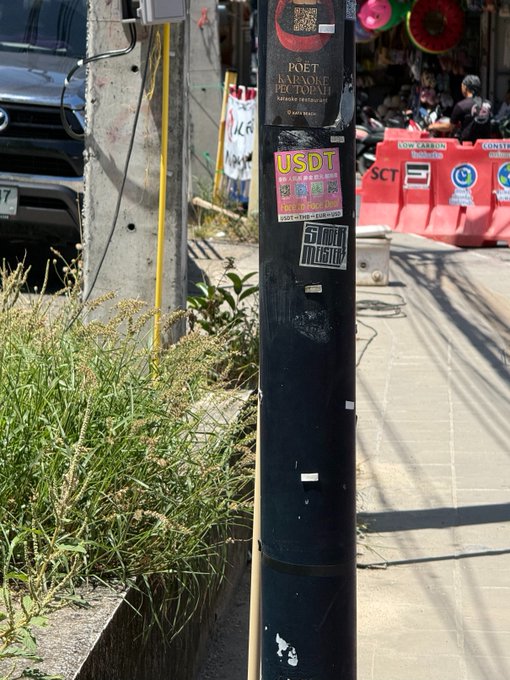 A black telephone pole in what appears to be Thailand is plastered with modern flyers, including a bright pink one advertising "USDT" purchases and QR codes, alongside others mentioning "POET รับลงทะเบียน" and Thai restaurant signage in the background. The image illustrates the post's contrast between old-school pole flyers offering unconventional services and today's crypto-focused advertisements.