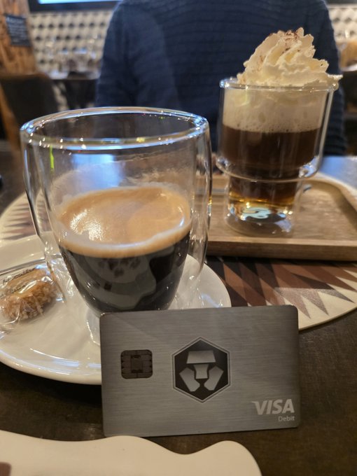 A Visa debit card with a metal finish sits in front of two glass cups of coffee, one topped with whipped cream, on a café table with a decorative patterned plate.