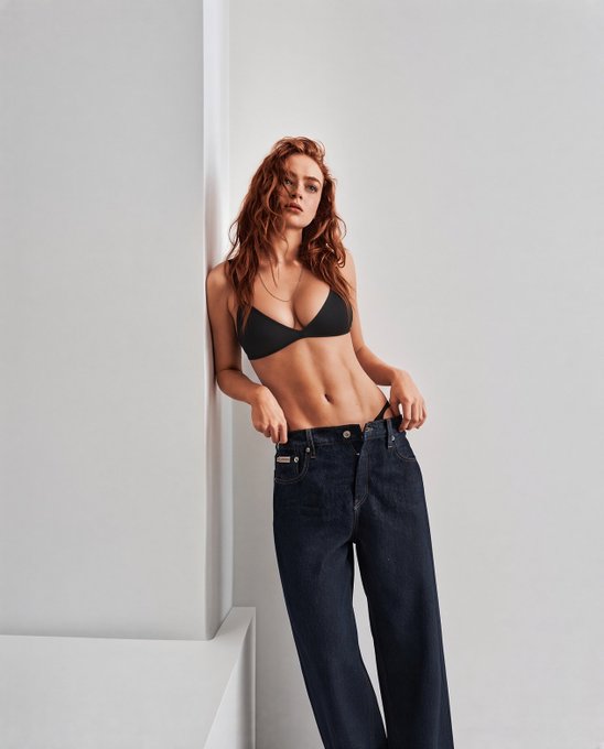 A woman with long red hair posing in a black bra and unbuttoned dark jeans against a white wall.