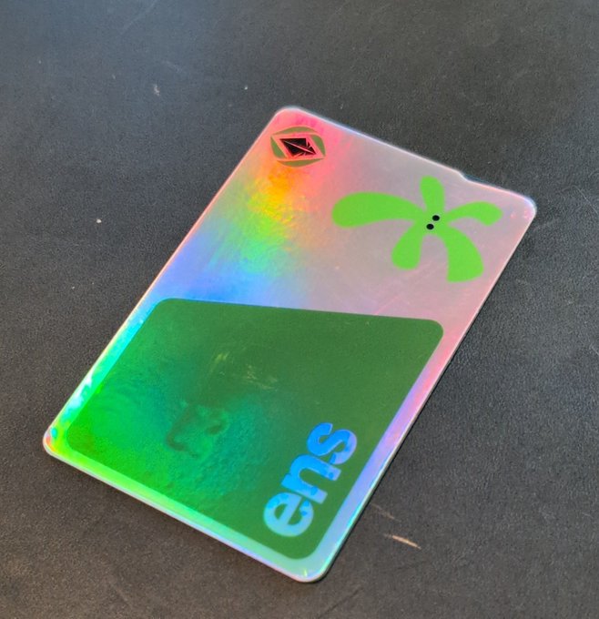 A holographic NFT card with 'eπs' text and a green splash graphic rests on a dark surface.