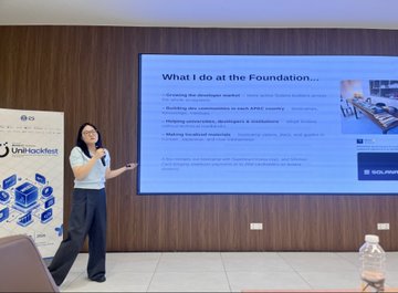 A woman presents on stage at a Unirackfest event, gesturing toward a slide titled "What I do at the Foundation..." which lists responsibilities including expanding the developer ecosystem, building new communities, and helping communities/institutions. The presentation, connected to a discussion about Solana, showcases her role and engagement within the blockchain community.