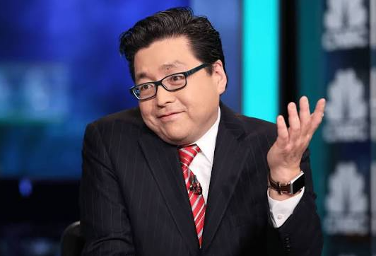 A portrait of a smiling Asian man wearing glasses, a dark pinstripe suit, and a red striped tie, gesturing with his hand during what appears to be a TV interview.