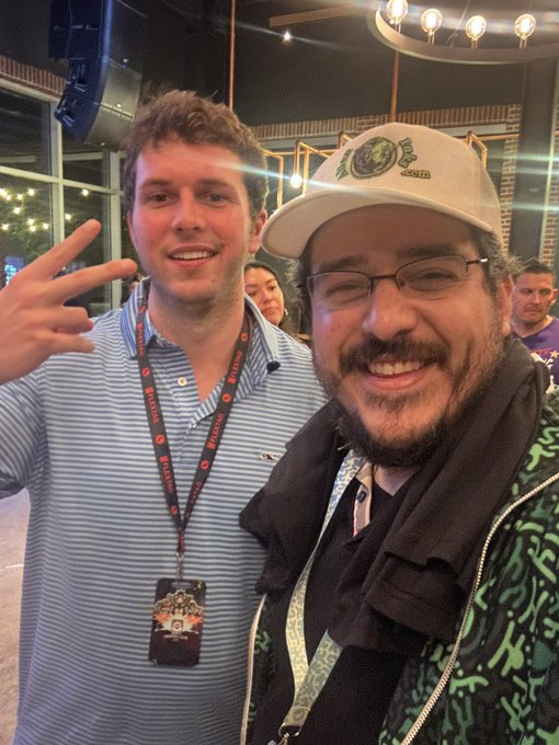 Two men smiling and posing for a photo at what appears to be an indoor event or bar, with one wearing a baseball cap, glasses, and a patterned jacket, while the other wears a blue striped shirt and a lanyard with a 'Crunchyroll Expo' badge.
