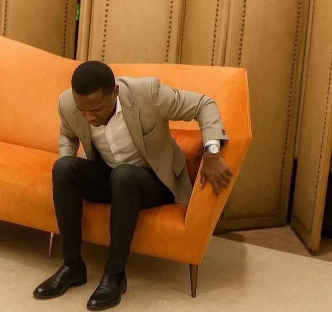 A well-dressed man in a beige blazer slumps forward on an orange couch with his head bowed, embodying the defeated "e go be na" (Nigerian slang for "it will be well") sentiment of reluctantly accepting an uncertain prediction on Polymarket.

No visible text, numbers, or names appear in the image.