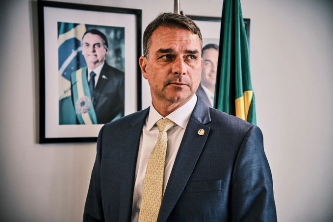 Flávio Bolsonaro stands in a formal suit with a Brazilian flag visible behind him and a framed portrait of himself on the wall, an image that visually anchors the post about his rising odds on Polymarket to become Brazil's next president, now tied with incumbent Lula at 50% and 46% respectively after climbing from 21.9% in July.
