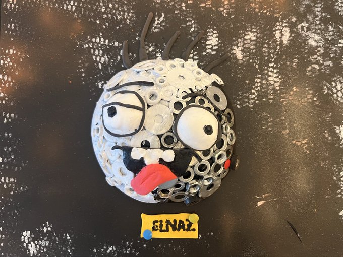 A child's craft project depicting a quirky face made from white buttons, googly eyes, and a red clay tongue on a black sparkly background, labeled with a yellow tag reading 'ELNAZ'.