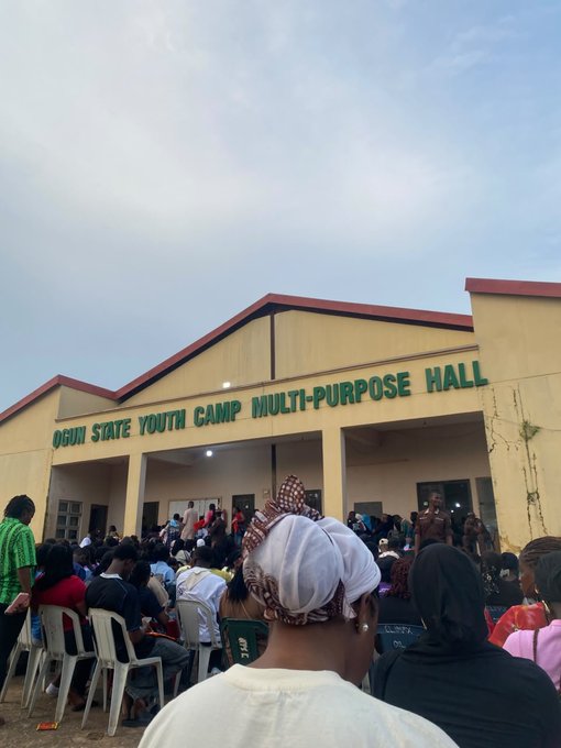 An outdoor gathering of people seated in chairs facing a building labeled 'OGUN STATE YOUTH CAMP MULTI-PURPOSE HALL' under a cloudy sky.