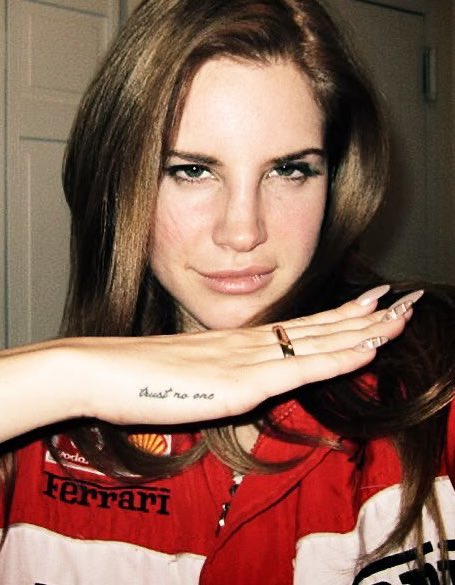 A woman wearing a red Ferrari racing shirt poses with her hand near her face, showing off a 'trust no one' wrist tattoo and long manicured nails.