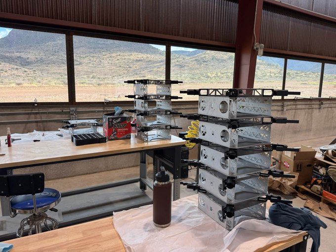 A robotics workspace with stacks of metal frames and mechanical components on wooden tables, featuring a scenic mountain view through large windows.