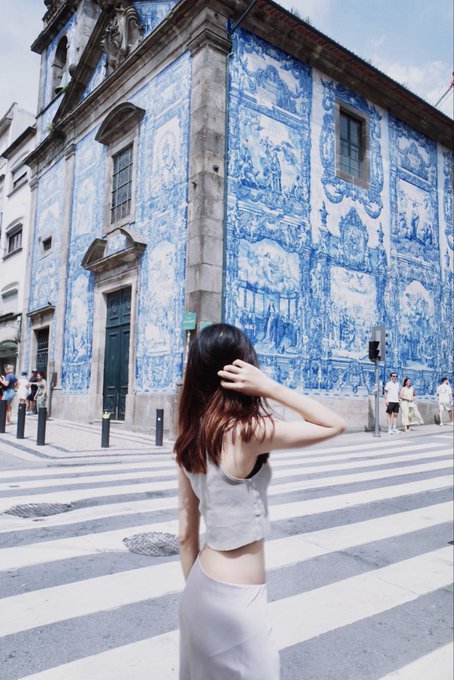 A young woman with long brown hair stands on a crosswalk, gazing up at a striking historic building covered in intricate blue and white azulejo tiles in what appears to be Porto, Portugal. The caption highlights how she balances rural and urban travel while testing the @ether_fi @BitgetWalletCN card along a scenic pilgrimage route, perfectly capturing the unexpected blend of historic European city exploration with modern crypto adoption.